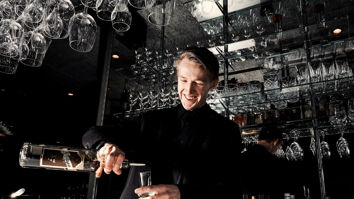 Lachender Barkeeper gießt an der Bar ein Getränk ein, darüber das Regal mit hängenden Gläsern
