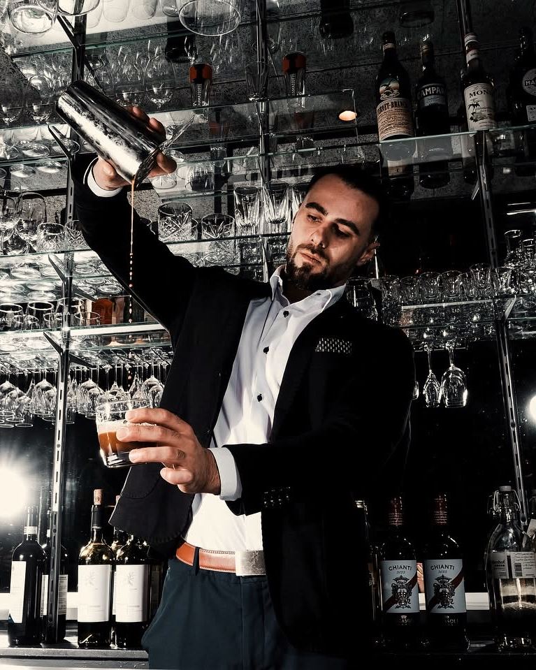 Barkeeper in Weste schüttelt hinter der Bar einen Cocktail, darüber das Gläserregal