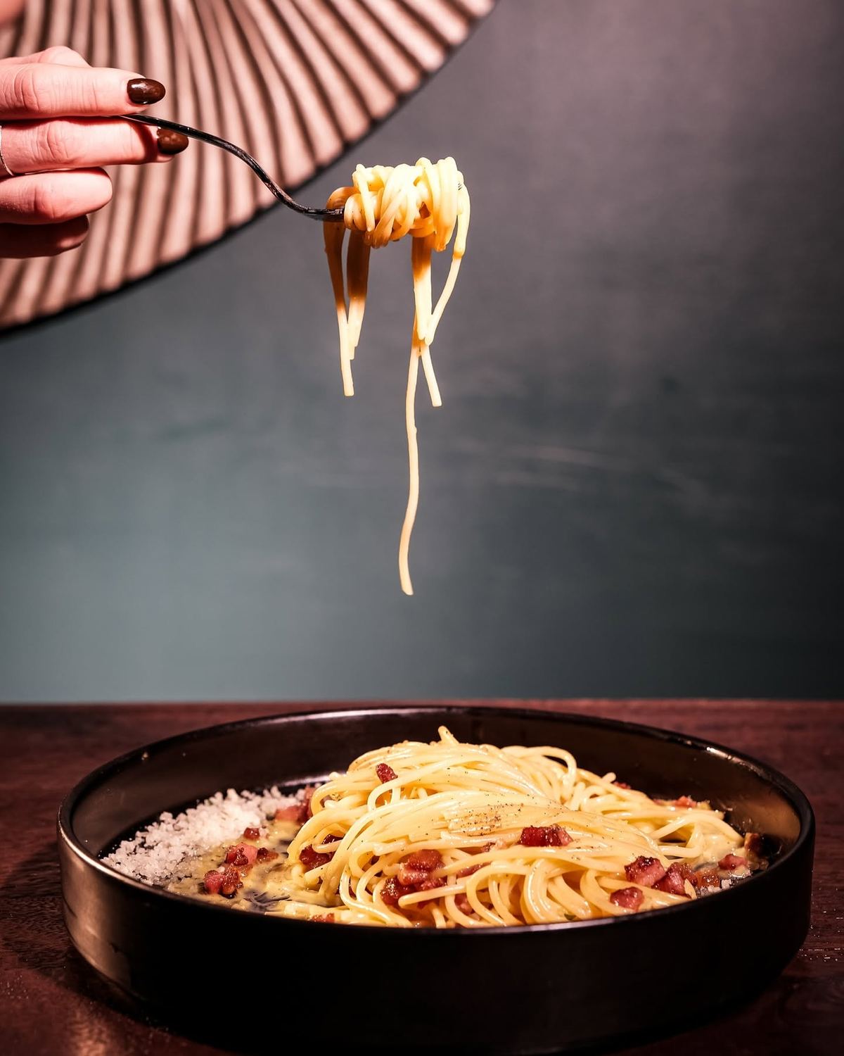 Eine Gabel hebt Spaghetti Carbonara aus einer dunklen Schale, dahinter die petrolgrüne Wand