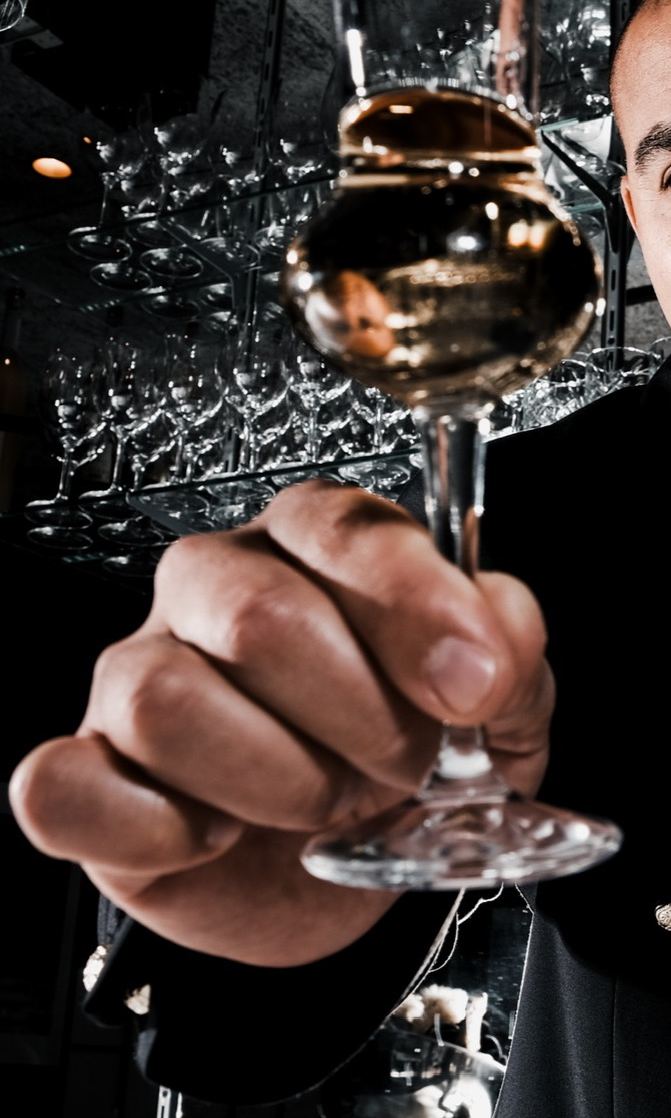 Hand hält ein Stielglas mit hellem Getränk vor dem Gläserregal der Bar